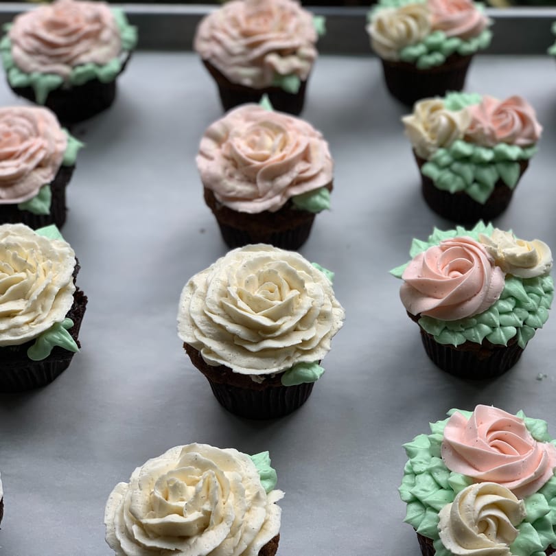 Chocolate cupcakes with pink and white rose-shaped frosting and green leaves on a baking sheet.